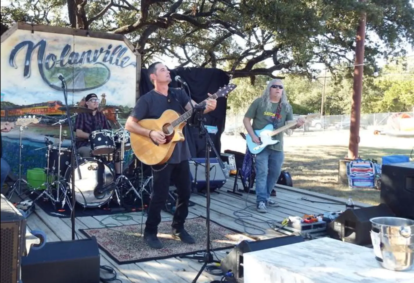 Local band performing on an outdoor stage at a Nolanville community event, with musicians playing guitar and drums under shaded trees.